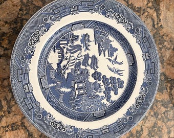 May include: A round ceramic plate featuring a classic blue and white Willow pattern. The design depicts a detailed landscape with a pagoda, a bridge, willow trees, and two birds in flight, surrounded by a wide, intricate geometric border.