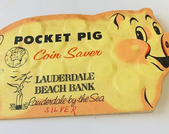 May include: Vintage Pocket Pig coin saver in the shape of a pig. The yellow and orange cardboard coin bank features the text "Pocket Pig Coin Saver" and "Lauderdale Beach Bank". The pig has a smiling face and coin-sized holes.