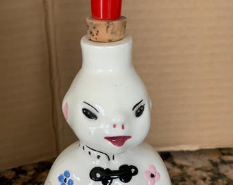 May include: A white ceramic bottle with a face design, featuring a red stopper and a cork. The bottle is decorated with blue and pink floral accents and a black bow tie. The bottle has a unique, vintage aesthetic.