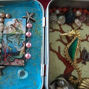 May include: A vintage tin box with two compartments. The left compartment features a blue background with a mermaid illustration and pink and white beads. The right compartment has a green and gold seahorse with a green stone on a coral background with orange beads.