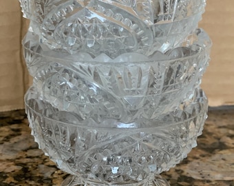 May include: A stack of three clear glass bowls featuring a detailed pressed pattern with geometric shapes and a scalloped rim. Each bowl has a circular base and a textured exterior surface that reflects light through the transparent glass material.