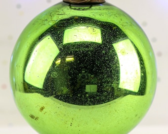 May include: A large, round, green Christmas ornament with a metallic, antique-style cap. The glass surface reflects light, creating a shiny appearance. The ornament has a vintage, distressed look.