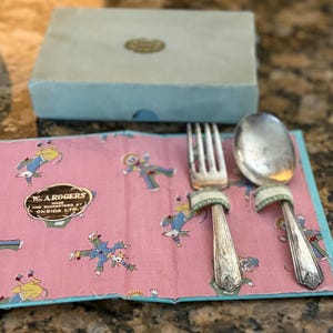 May include: Vintage silverware set with a fork and spoon, resting on a pink cloth with clown illustrations. The cloth is bordered in blue and features the text "W.A. ROGERS". A light blue box is in the background.