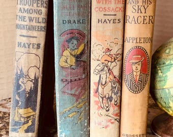 May include: Four vintage hardcover books standing upright. The spines read: The Boy Troopers Among the Wild Mountaineers by Hayes, The Boy Allies with the Great Powers by Drake, The Boy Allies with the Cossacks by Hayes, and Tom Swift and His Sky Racer by Appleton.