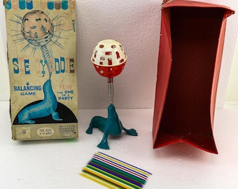 May include: Vintage "See-Yodle" balancing game by Milton Bradley. The game features a blue seal balancing a red and white ball on a spring, a red container, a box with game instructions, and colorful sticks. For ages 8 to adult.