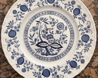 May include: A round white ceramic plate featuring a blue floral and fruit pattern. The center displays a detailed botanical motif enclosed by a decorative border, while the rim is adorned with repeating blue fruit and leaf illustrations.