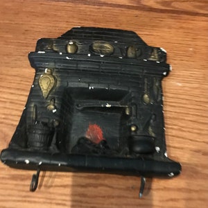May include: Black and white ceramic fireplace wall decor with a red painted fire and gold accents. The fireplace has two hooks on the bottom.