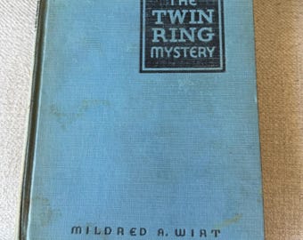 May include: A vintage book titled "The Twin Ring Mystery" by Mildred A. Wirt. The book has a light blue cover with the title in a black rectangular box. The author's name is printed in black at the bottom.