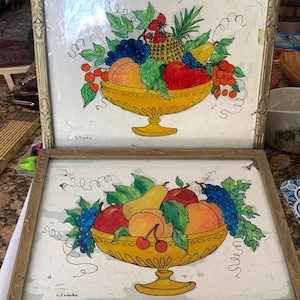 May include: Two framed pictures of fruit bowls with a white background. The fruit bowls are yellow and filled with fruit including peaches, pears, grapes, cherries, and a pineapple. The pictures are signed "L. Trinks".