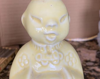 May include: A vintage yellow ceramic salt shaker in the shape of a person. The shaker has a metal top with holes and a green base. The figure has a serene expression and is wearing a decorative collar.