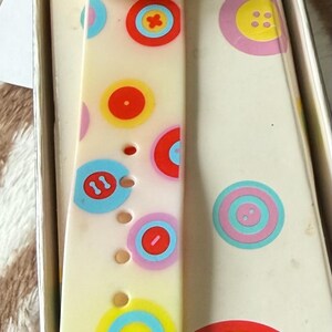 May include: A vintage Avon Button Watch in its original box. The watch features a cream-colored strap adorned with colorful button designs. The watch face is yellow and blue. The box has the words "AVON BUTTON WATCH".