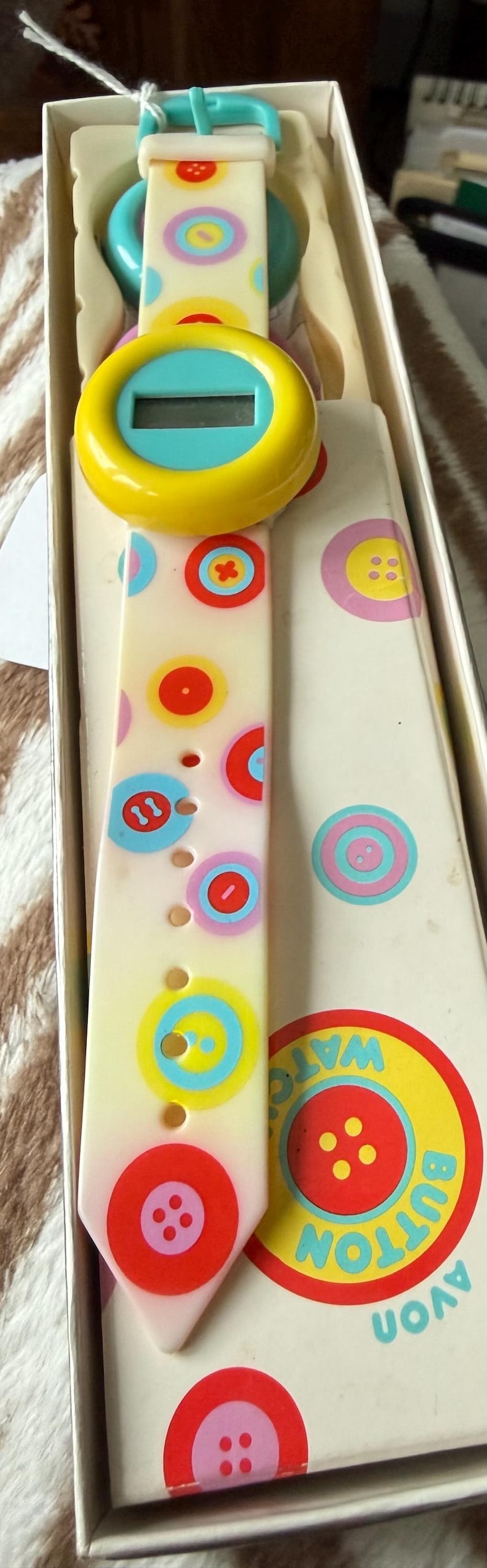 May include: A vintage Avon Button Watch in its original box. The watch features a cream-colored strap adorned with colorful button designs. The watch face is yellow and blue. The box has the words "AVON BUTTON WATCH".