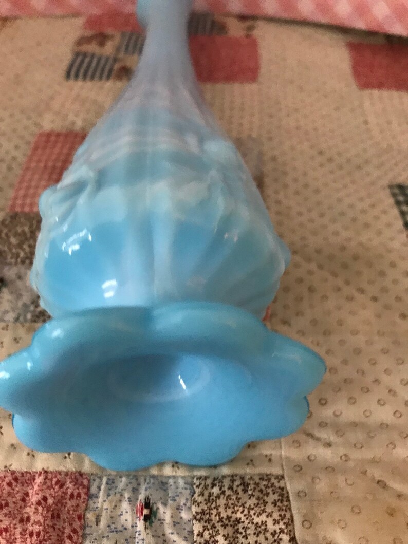 Puede incluir: Un jarr&oacute;n de cristal azul claro con base acampanada y cuello largo y delgado. El jarr&oacute;n tiene un dise&ntilde;o con volantes y texturizado y un acabado suave y opalino. El jarr&oacute;n est&aacute; sobre una tela estampada.
