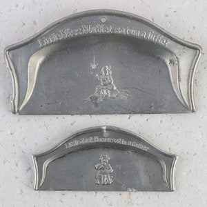 May include: Two vintage metal cookie cutters, one shaped like a girl sitting on a hutch with the text "Little Miss Muffet sat on a hutch", and the other shaped like a girl sitting in a corner with the text "Little Jack Horner sat in a corner".