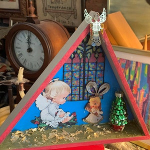 May include: A triangular wooden box with a red and green trim. The box is decorated with a stained glass window, a small angel figurine, and a miniature Christmas tree. The box is filled with gold glitter and has a blue background.