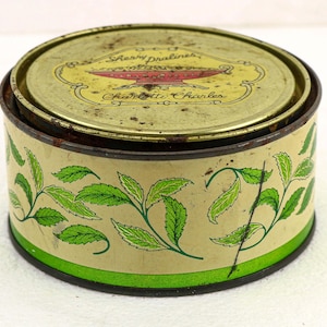 May include: Vintage round tin with a lid, featuring a cream-colored base and a green and gold design. The sides display green leaf patterns, while the lid has text and a red and gold emblem. The tin is slightly worn.