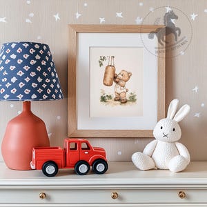 May include: A framed illustration of a brown bear wearing boxing gloves and punching a hanging punching bag. The illustration is set against a light brown background with green foliage. The frame is made of light wood.