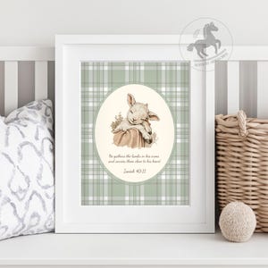 May include: A framed print featuring a sleeping lamb illustration in a soft, neutral color palette. The background has a green and white plaid pattern. The text reads, "He gathers the lambs in his arms and carries them close to his heart. Isaiah 40:11."