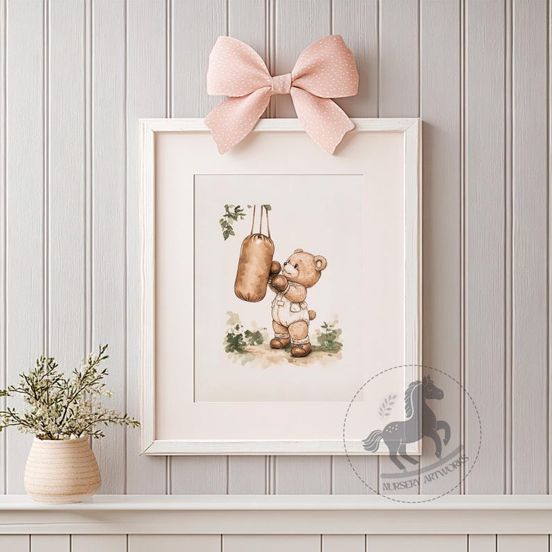 May include: A watercolor illustration of a brown bear wearing boxing gloves and punching a hanging punching bag. The bear is wearing a white onesie with a red belt. The illustration is framed in a white frame with a pink bow on top.