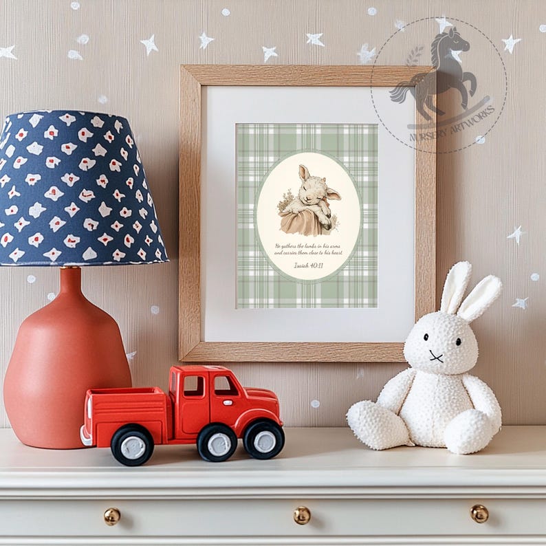 May include: A framed print with a lamb illustration on a green plaid background. The print includes text from Isaiah 40:11. A red toy truck, a white stuffed bunny, and a blue patterned lamp are on a white surface.