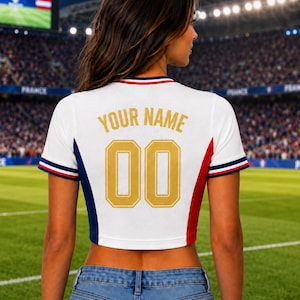 May include: White crop top with blue, red, and white trim, featuring "YOUR NAME" and the number "00" in gold lettering. The shirt has a sporty design, with a soccer field in the background.