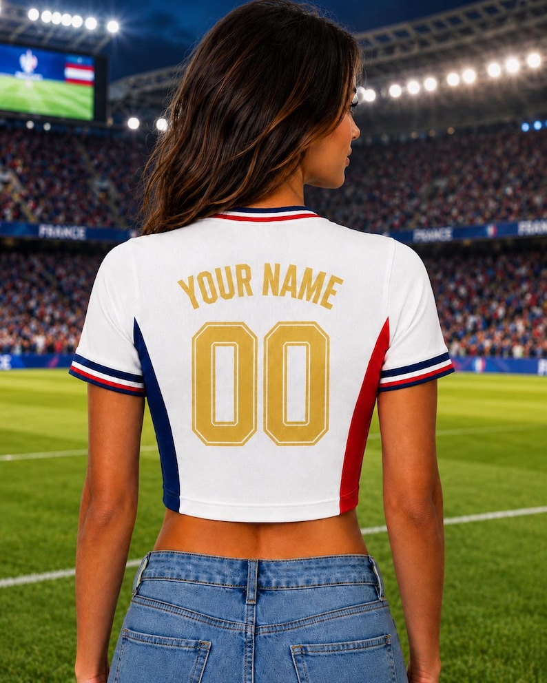 May include: White crop top with blue, red, and white trim, featuring "YOUR NAME" and the number "00" in gold lettering. The shirt has a sporty design, with a soccer field in the background.