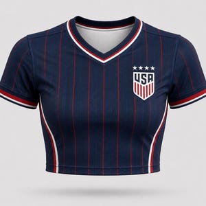 Front view of a women’s USA football away crop top in dark navy with thin red pinstripes, subtle panel details, layered collar and sleeve trims in red, white, and navy, and a shield-style USA crest on the chest