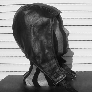 May include: A black leather hooded hat with a strap and buckle closure. The hat is worn on a mannequin head.