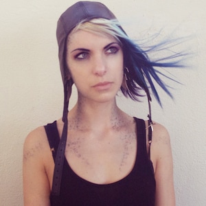 Può includere: Una donna con capelli blu e biondi indossa una canotta nera e un cappello da aviatore in pelle marrone con cinturini.