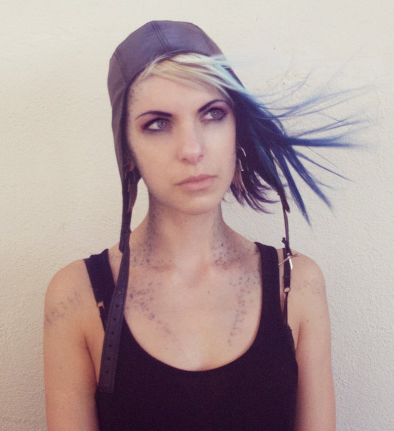 May include: A woman with blue and blonde hair is wearing a black tank top and a brown leather aviator hat with straps.