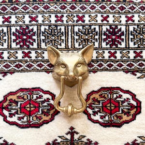 May include: A brass fox head door knocker with a detailed design. The knocker features a loop handle and is mounted on a decorative plate. The fox head is centered, with the loop handle hanging below. The background is a patterned rug with red, black, and beige colors.