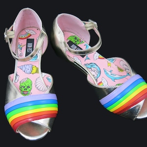 May include: A pair of gold sandals with a rainbow trim. The sandals have a pink insole with a whimsical design featuring a unicorn, ice cream cone, seashells, and a monster.