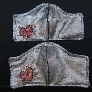 May include: Two iridescent silver face masks with black elastic ear loops. Each mask has a pink heart embroidered with safety pins.