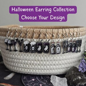 Könnte beinhalten: Eine Sammlung von Halloween-Ohrringen aus Acryl, die an einem geflochtenen Korb hängen. Die Motive umfassen Weingläser, Bierkrüge mit Totenköpfen, Geister-Getränkedosen und Giftfläschchen. Der Text lautet: Halloween Earring Collection Choose Your Design.