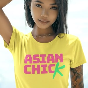 以下が含まれることがあります： ピンクと緑で「ASIAN CHIC」と書かれた黄色のTシャツ。クルーネックのスタイルで、柔らかい素材のようです。モデルはネックレスをしており、腕にタトゥーがあります。