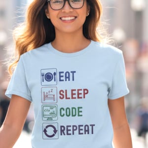 May include: Pale blue t-shirt featuring the text "EAT SLEEP CODE REPEAT" in a vertical arrangement. The words are in blue, red, green, and purple. The shirt has a round neck and short sleeves.
