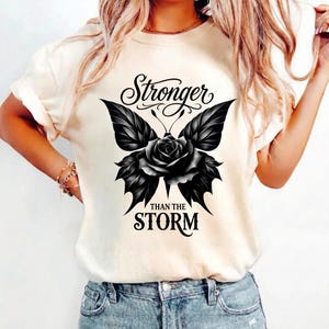 Może przedstawiać: Kremowa koszulka z czarnym motylem i różą pośrodku. Skrzydła motyla mają wzory przypominające liście. Tekst "Stronger than the Storm" jest wygięty nad i pod motylem.