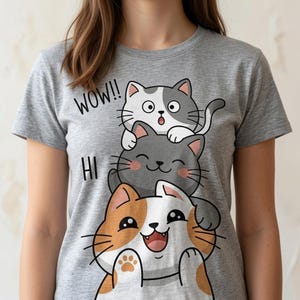 May include: A heather gray short-sleeve t-shirt featuring a graphic of three stacked cats. The top cat is white and gray with the text WOW!!, the middle cat is dark gray, and the bottom cat is orange and white with the text HI.