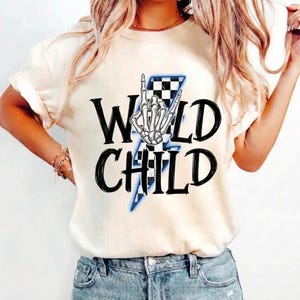 May include: A cream-coloured t-shirt featuring the text "WILD CHILD" in bold black lettering. A skeleton hand in a rock and roll pose is above a blue and black chequered lightning bolt graphic.