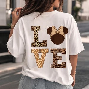 May include: A white short-sleeve t-shirt featuring a large graphic on the back. The design displays the word LOVE in block letters, where the L, V, and E have a leopard and giraffe print pattern, and the O is replaced by a brown mouse-ear silhouette.
