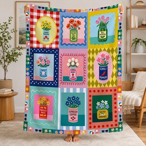 May include: A rectangular throw blanket featuring a grid of nine colorful illustrations of flowers in various containers, including cans and jars labeled Cherry Tomatoes, Olive Oil, Dill Pickles, Sweet Bloom, Berry Jam, Chili Peppers, Farfalle Pasta, Espresso Coffee, and Salted Sardines.