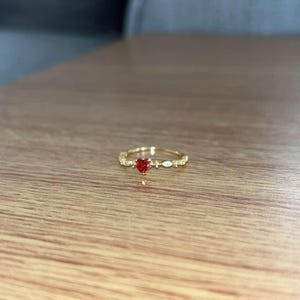 Op de afbeelding: Een gouden ring met een kleine rode hartvormige steen en heldere stenen gezet in een delicate band.