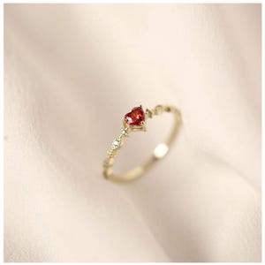 May include: A delicate gold ring featuring a small red heart-shaped gemstone. The ring has a dainty design with small accents.