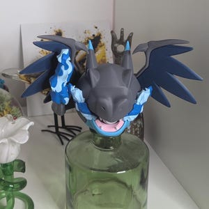 Peut inclure: Figurine de dragon gris et bleu avec des ailes déployées, posée sur une bouteille en verre verte. Le dragon a des flammes et des accents bleus, avec un visage détaillé et une bouche ouverte. Les ailes sont bleu foncé.