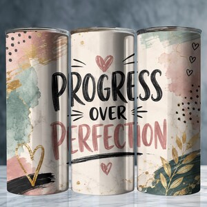 May include: Three cylindrical tumblers featuring a cream background with abstract watercolor splashes in sage green, dusty pink, and gold. The center tumbler displays the black and pink text "PROGRESS OVER PERFECTION" with small heart icons and gold leaf accents.