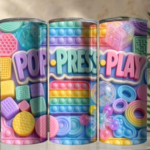 May include: A cylindrical tumbler featuring a colorful pattern of various sensory fidget toys in pastel shades of pink, blue, yellow, and green. The design includes the text POP, PRESS, and PLAY in large, raised-style letters across the center.