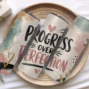 May include: Three cylindrical tumblers featuring a cream background with watercolor-style splashes in sage green, dusty pink, and black. The text "PROGRESS OVER PERFECTION" is printed in black and pink, accented by small hand-drawn hearts and gold lines.