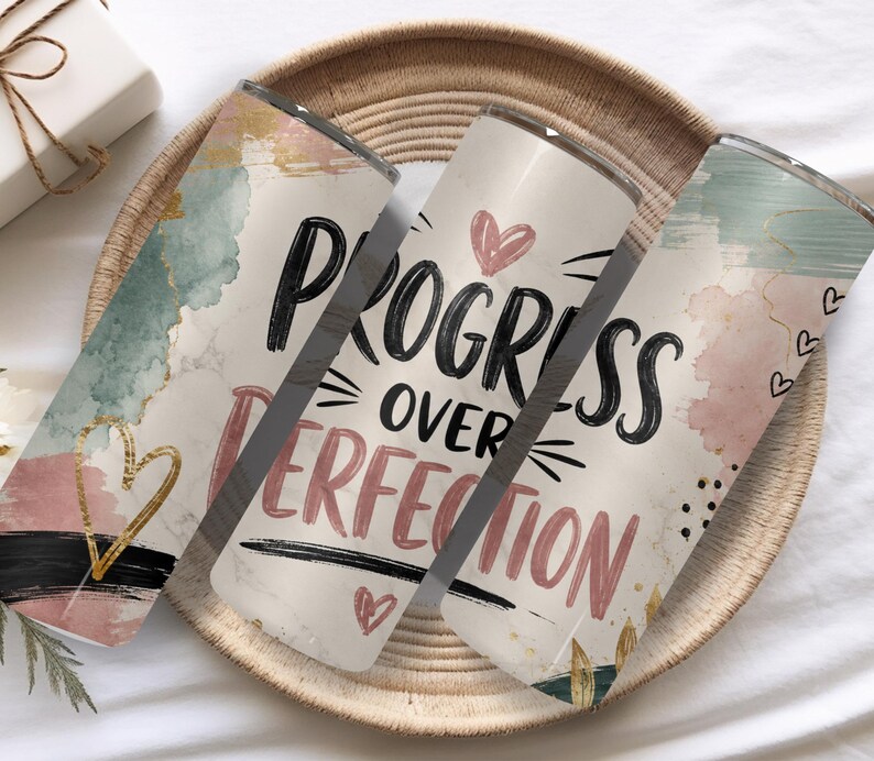 May include: Three cylindrical tumblers featuring a cream background with watercolor-style splashes in sage green, dusty pink, and black. The text "PROGRESS OVER PERFECTION" is printed in black and pink, accented by small hand-drawn hearts and gold lines.