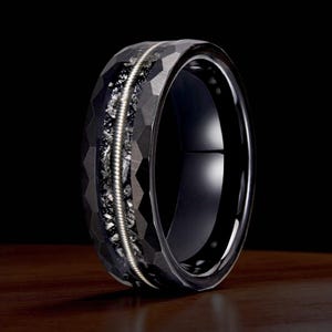May include: A black ring with a hammered finish and a central inlay of silver-coloured metal and small, sparkling stones. The ring has a polished, glossy interior and sits on a wooden surface.