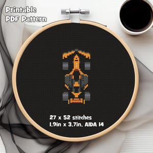 May include: A cross-stitch pattern of a Formula 1 race car in orange, grey, and black, displayed in a wooden embroidery hoop. The text "Printable PDF Pattern" is at the top. The pattern is 4.8cm x 9.4cm and has 27 x 52 stitches.